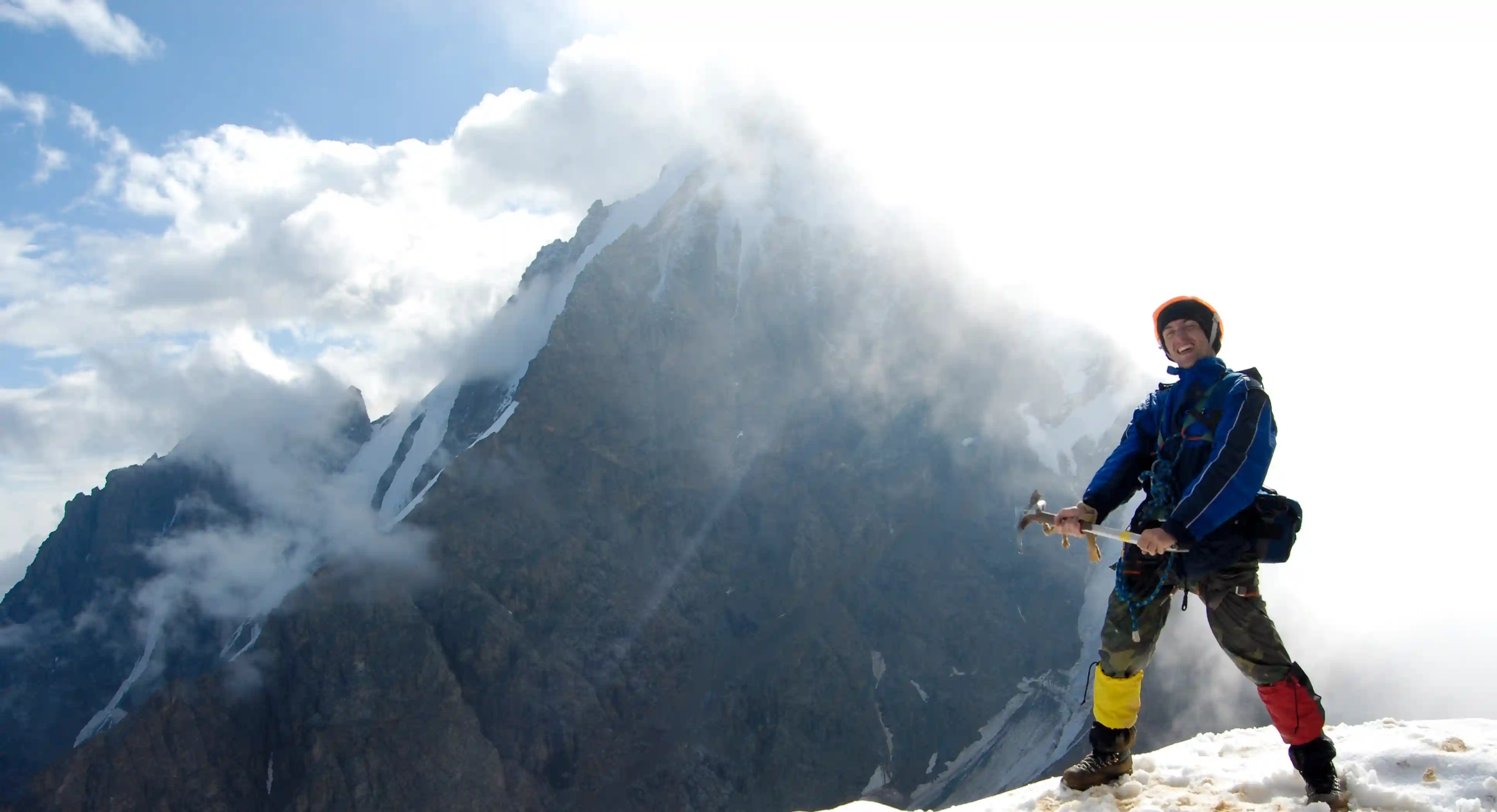 A mountaineer in a red jacket, yellow helmet, and blue backpack uses an ice axe and crampons to scale a steep, sunlit glacier against a clear blue sky.